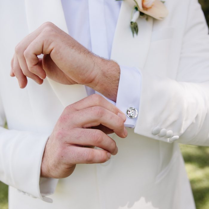 A person in a white suit jacket and black bow tie adjusts a cufflink on their shirt sleeve, with a light colored boutonnière on the lapel.