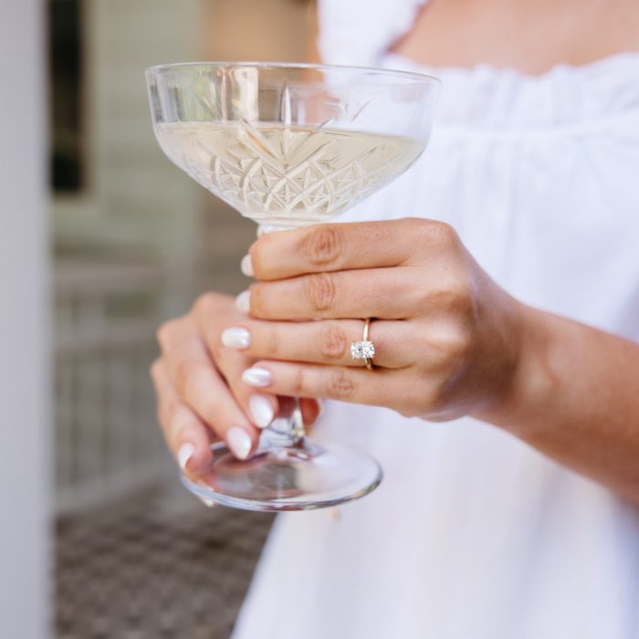 A person in a white top holds a coupe glass with a light colored beverage, displaying a diamond ring on their left hand.
