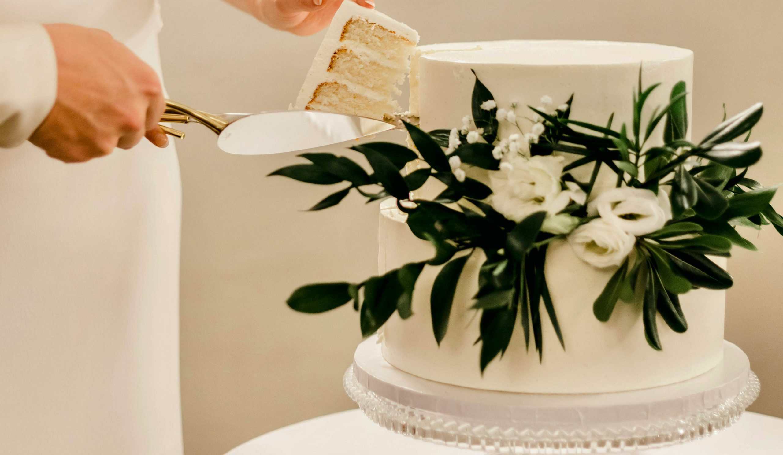 A person cuts a slice from a white, two tier cake decorated with green leaves and white flowers on a clear cake stand.
