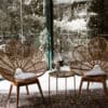 Two rattan chairs with petal-shaped backs face each other in a sunlit room, perfect for event hire. A Side Table l Round Gold adorned with a floral arrangement stands between them on a patterned rug, with greenery visible outside, encapsulating the essence of a Gold Coast wedding.