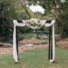 An arbour adorned with white chiffon and flowers is set up on a grassy area by a small stream and trees, available through Gold Coast wedding hire.