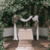 Experience an enchanting outdoor event with our "Arbour l Dark Timber," featuring a wedding altar with a table draped in lace beneath this elegant arch. Adorned with blooming flowers, it is set against the scenic backdrop of lush greenery, creating the perfect ambiance for your special occasion. Available exclusively through our event hire services.