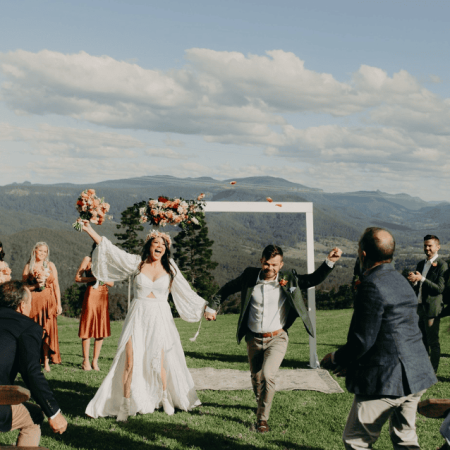 A bride and groom celebrate their wedding outdoors, surrounded by a joyful wedding party. The stunning event unfolds against a backdrop of majestic mountains and a clear sky, featuring the elegant White Timber Arbour as a central piece of the ceremony.
