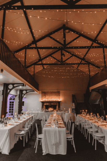 The hall, ideal for a Gold Coast wedding event, showcases long tables adorned with off white cheesecloth table runners and chairs. The ceiling, embellished with string lights, creates a warm ambiance that enhances the celebratory atmosphere.