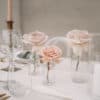A beautifully arranged table adorned with empty glasses, elegant candlesticks, and light pink roses in the Floral Vase l Glass Bud captures the essence of a dreamy wedding event. Perfect for hire, the setting is completed with exquisite tableware that adds a touch of sophistication.