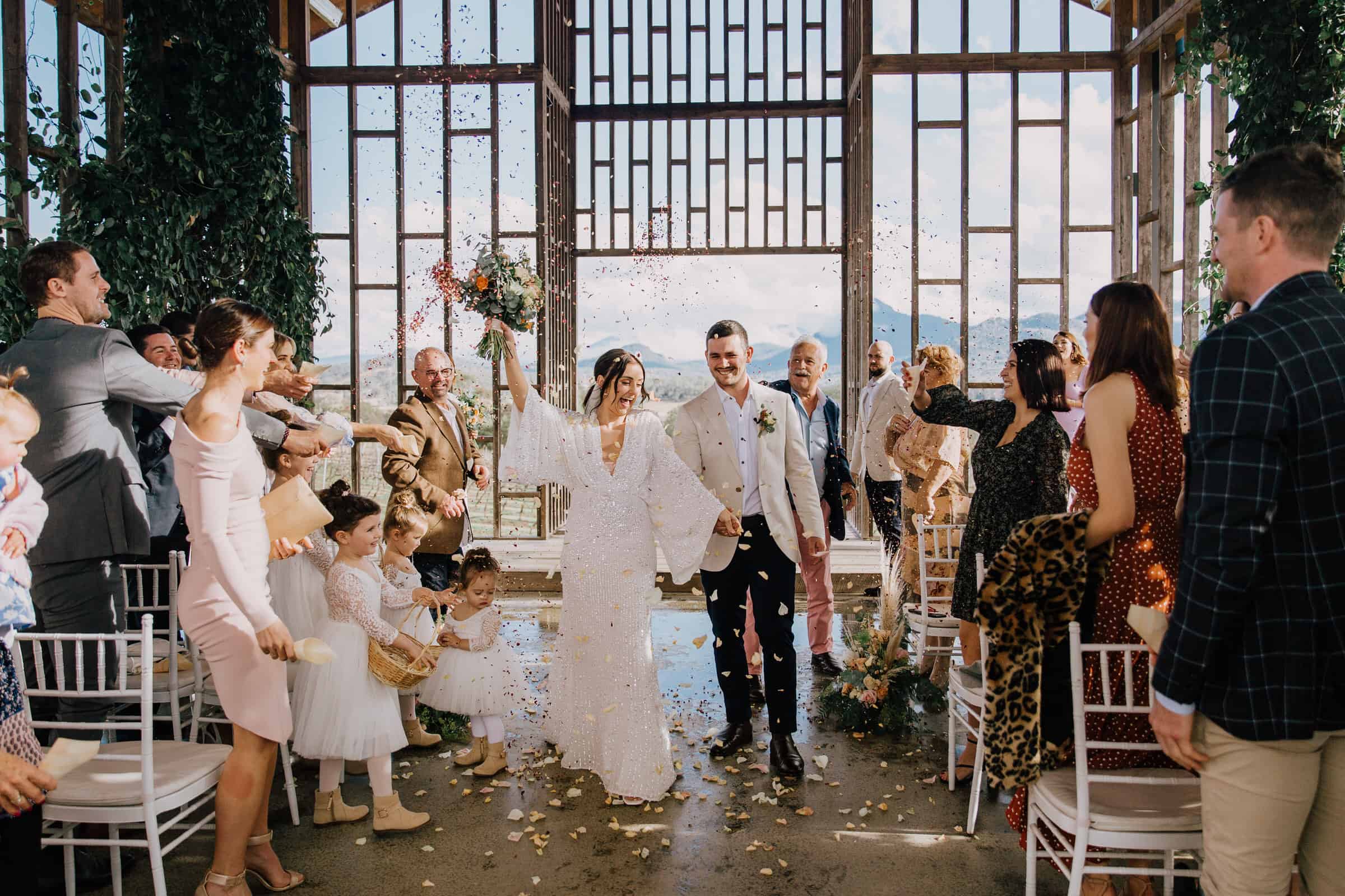 Bride and groom walk down the aisle under a shower of flower petals, surrounded by cheering guests inside a modern, sunlit venue, thanks to impeccable Gold Coast event hire.