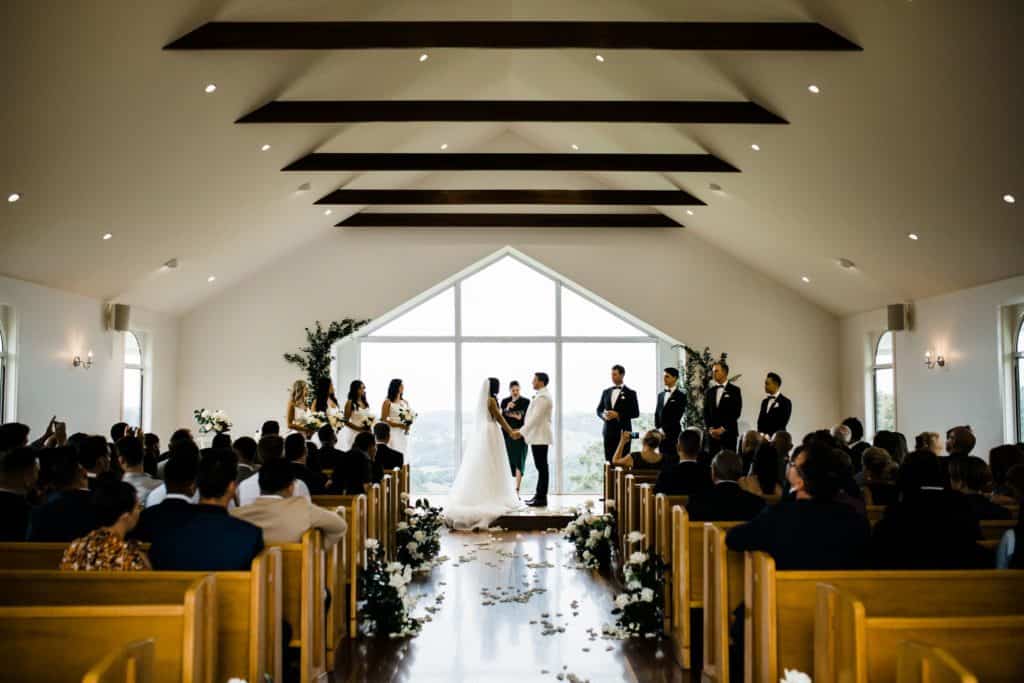 A couple stands at the altar in a church, holding hands. Bridesmaids and groomsmen are on either side, with guests seated in wooden pews. The large window behind them showcases a scenic view, beautifully framed with decor from the finest Gold Coast wedding hire service, elevating the event.
