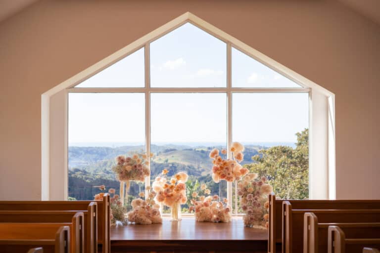 The chapel interior, perfect for a Gold Coast wedding hire, features a large window framing a scenic landscape. Wooden pews line the aisle, complemented by floral arrangements of soft-colored flowers that adorn the altar.