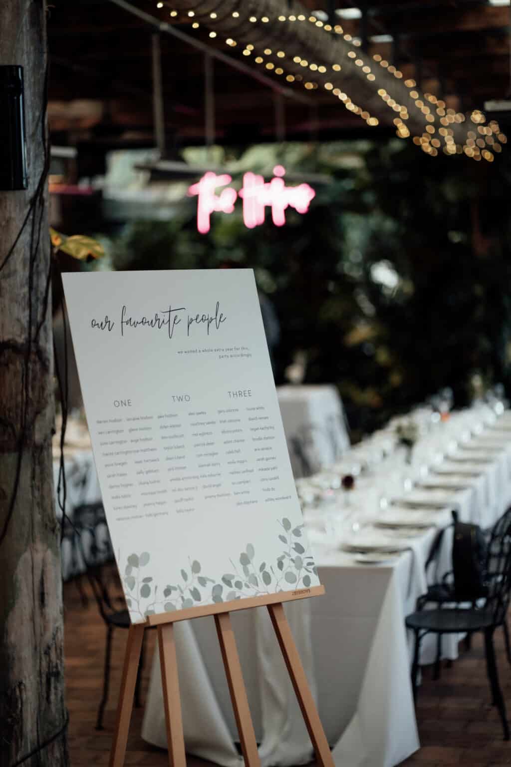 A beautifully curated wedding seating chart stands elegantly on an easel at the venue, provided by Gold Coast wedding hire. Tables are set perfectly, with string lights twinkling softly in the background, creating a magical atmosphere for this special event.