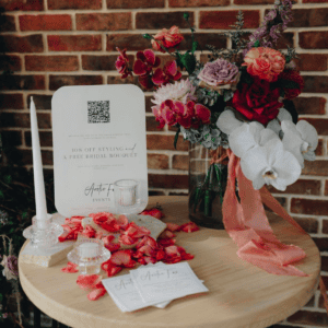 A table featuring a floral arrangement, business cards, and a QR code sign is elegantly set up for your wedding. Scattered rose petals accompany the 2-In-1 Candle Holder l Ava and decorations in front of a brick wall to complete the enchanting scene—perfect for an unforgettable Gold Coast hire event.