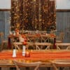 Wooden tables featuring Terracotta Cheesecloth runners, complemented by floral centerpieces and chairs, set in a room with rustic decor and string lights, create the perfect ambiance for your event. Experience the charm of Gold Coast wedding hire to transform any occasion into a memorable celebration.