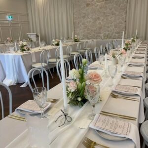 A long, elegantly set banquet table with white linens, floral centerpieces, Amelia Taper Candle Holders, and menus graces the bright room with a stone wall accent, epitomizing exquisite Gold Coast wedding hire.