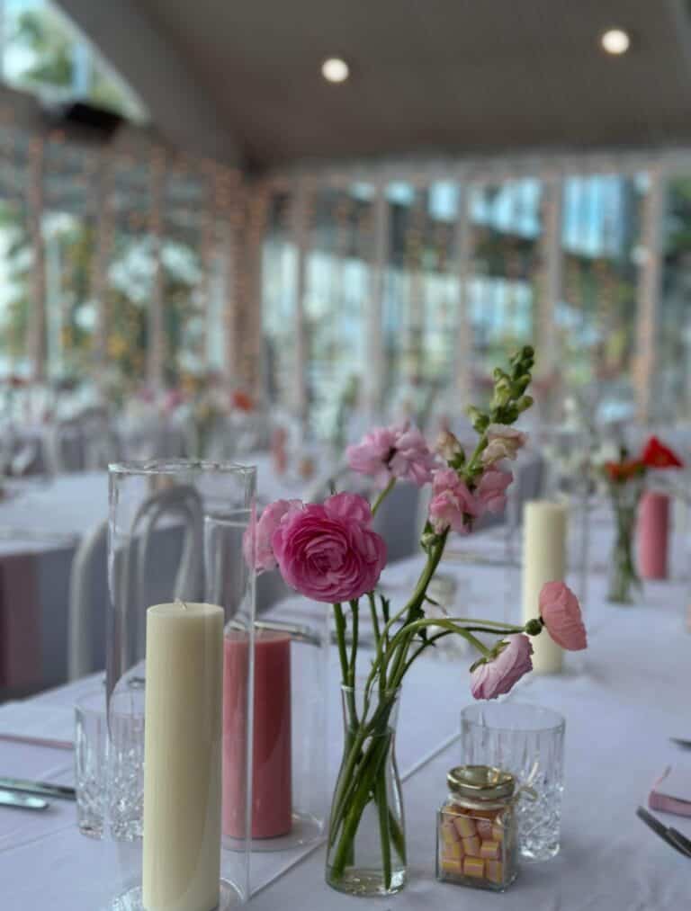 A wedding table setting with pink flowers in a Curvy Clear Bud floral vase, tall white candles, and glassware alongside a jar of candies. The blurred background features large windows and string lights, perfect for event hire to add an elegant touch.