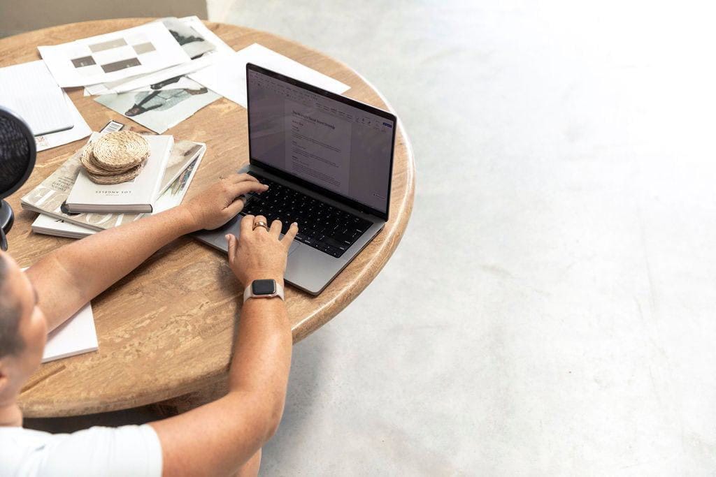 A person is typing on a laptop at a round wooden table, surrounded by scattered papers and notebooks, perhaps organizing details for an upcoming event. A watch on their wrist keeps them on schedule.