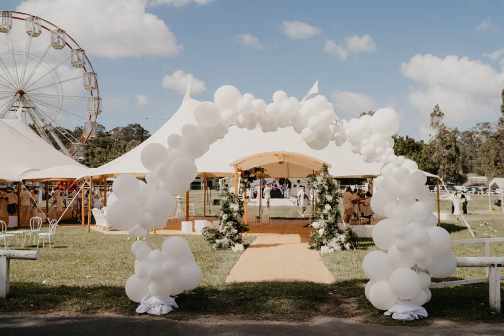The outdoor event setup features elegant white tents and a whimsical balloon arch on a lush grassy area, perfect for any wedding celebration. In the background, a Ferris wheel adds charm beneath the partly cloudy sky, showcasing the potential of Gold Coast wedding hire.