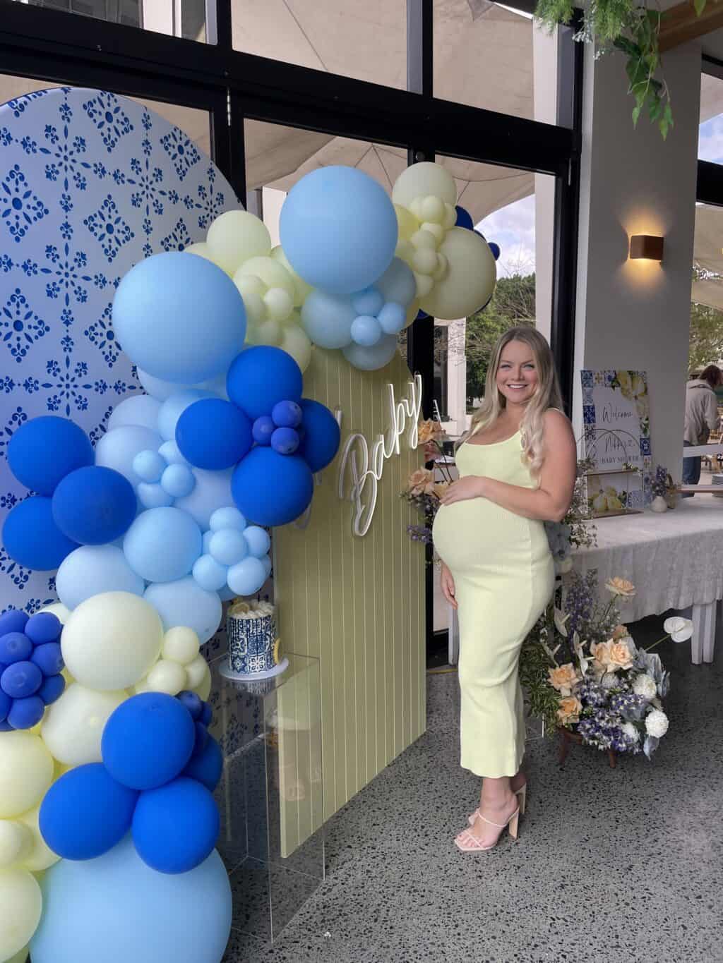 A pregnant woman in a light green dress stands by an elegant baby shower display, meticulously styled by a local event hire company. Surrounded by blue and white balloons, a "Baby" sign, and exquisite floral arrangements, she brings joy to this special Gold Coast event.