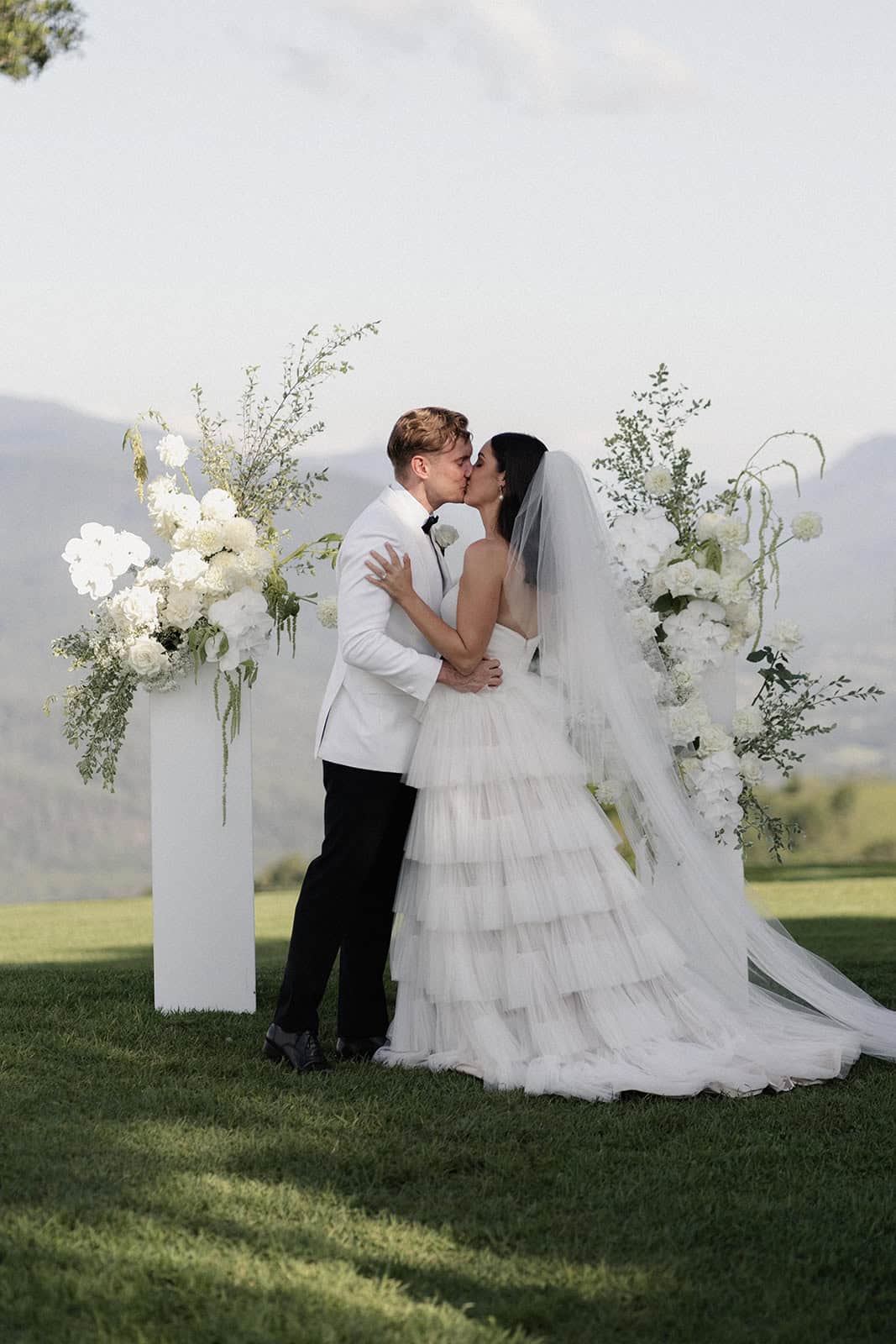 A couple kisses at an outdoor wedding ceremony, with floral arrangements and mountains in the background, creating a picture-perfect event for guests to cherish. For those planning their own unforgettable moment, finding the right event hire can transform any setting into a dreamscape.