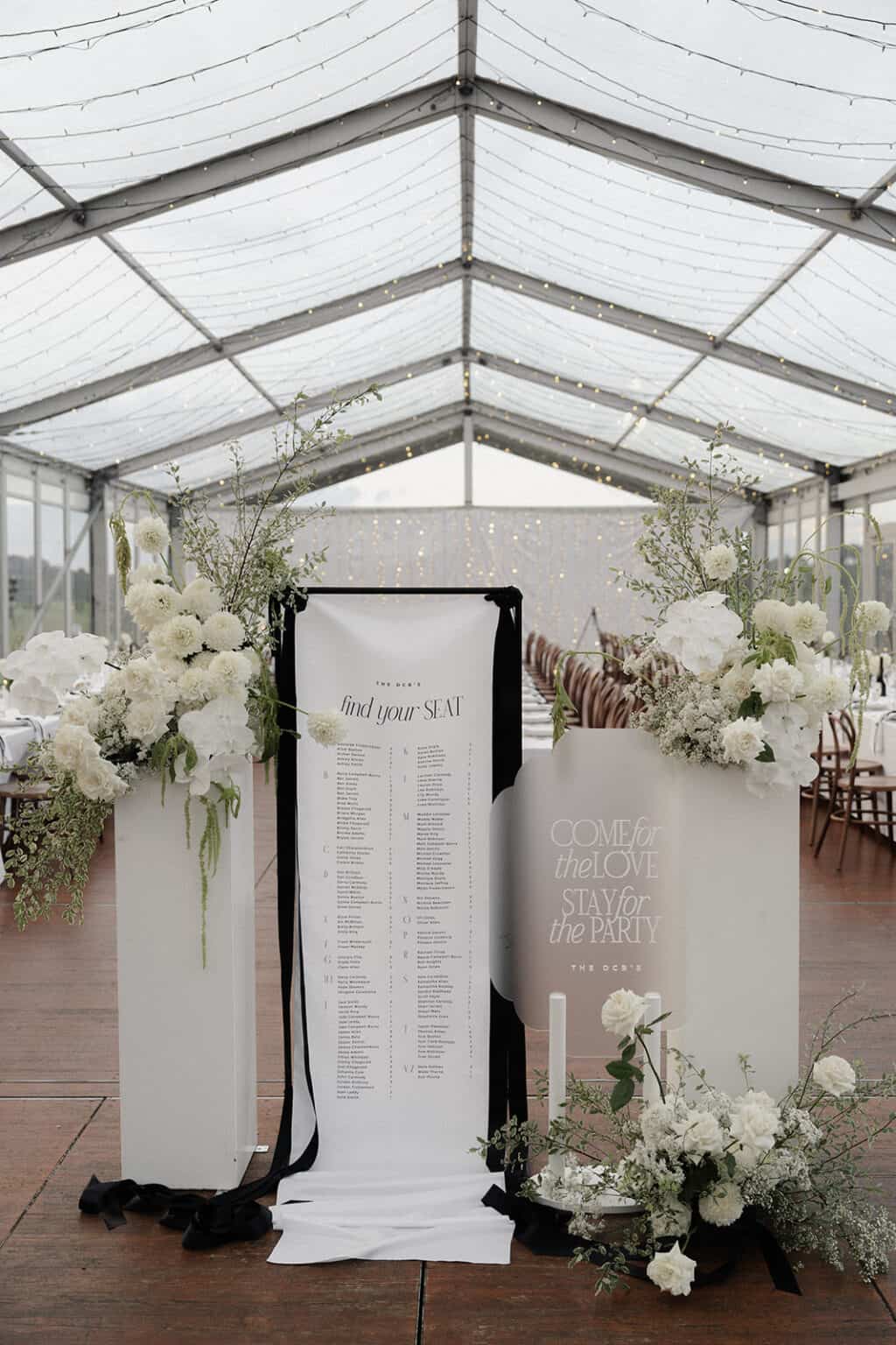 Wedding seating chart decorated with white flowers and greenery under a clear tent. Perfect for your Gold Coast hire needs, the setup includes tables and chairs elegantly arranged in the background, creating a dreamy wedding atmosphere.