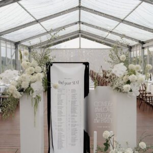 Wedding seating chart decorated with white flowers and greenery under a clear tent. Perfect for your Gold Coast hire needs, the setup includes tables and chairs elegantly arranged in the background, creating a dreamy wedding atmosphere.