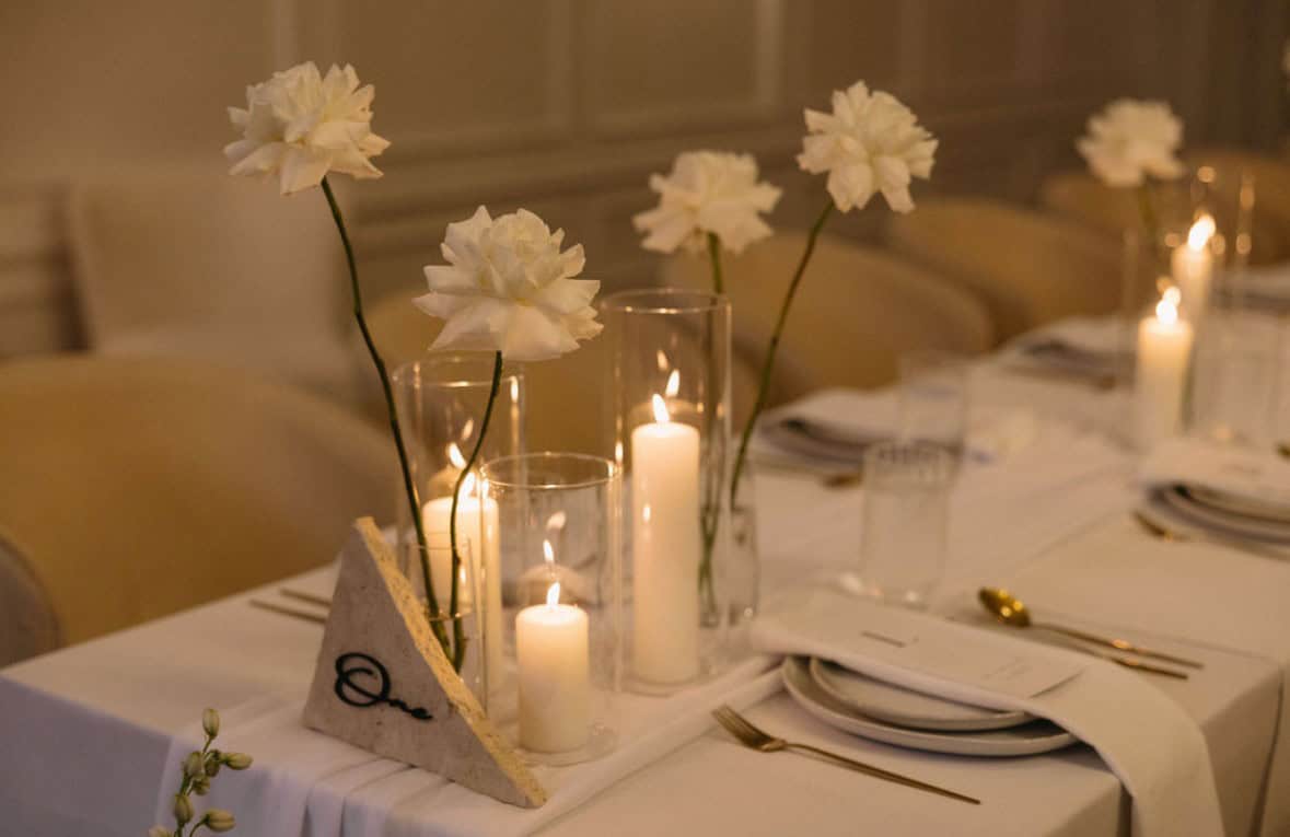 Create an elegant table setting for any event with white flowers in vases and lit candles. Neatly arranged plates, cutlery, and the sophisticated Table Numbers in Small Stone & Black add the perfect finishing touch, all available through our event hire service.