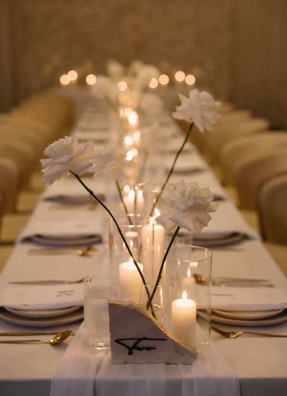 Elegant table setting featuring Table Numbers in Small Stone & Black, surrounded by white flowers, candles, and neatly arranged cutlery on a long table, perfect for a sophisticated wedding event.