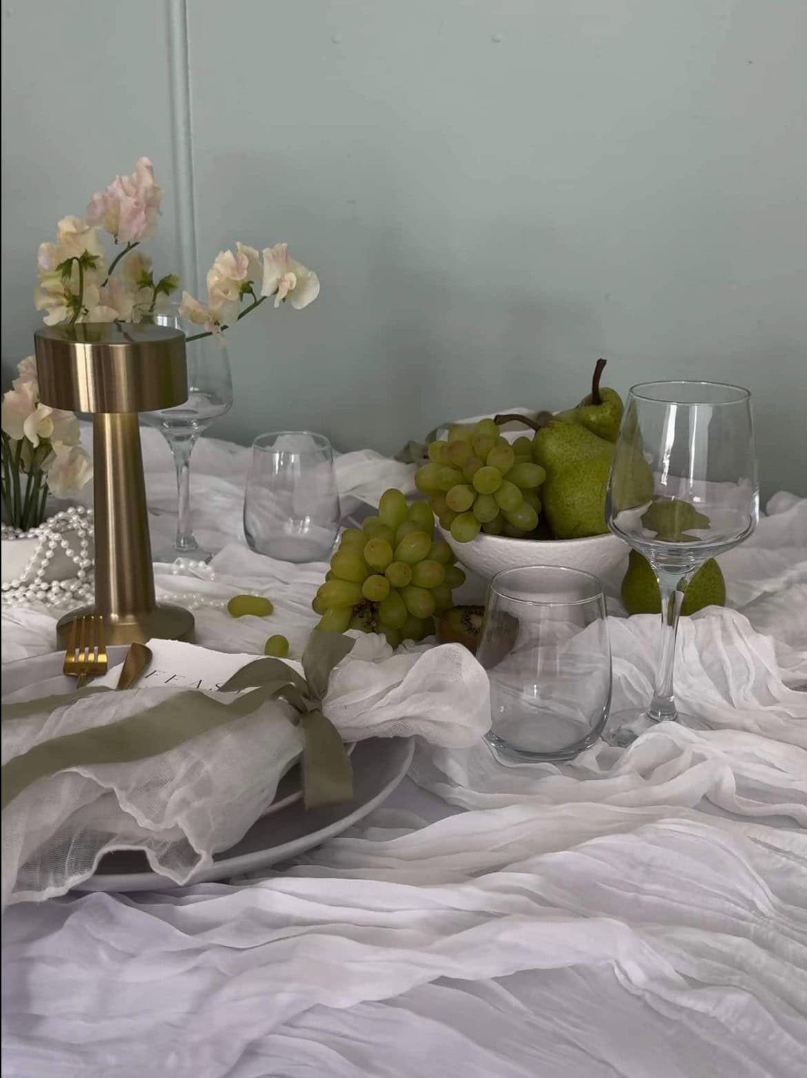 A neatly arranged table for a wedding is adorned with white flowers, grapes, pears, and elegant glassware on a white textured tablecloth, complemented by the refined touch of Napkin l White Gauze.