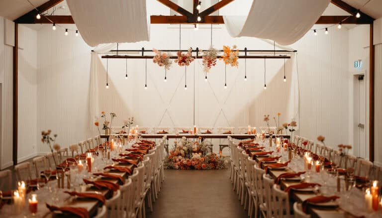 Long banquet tables with white chairs and rust colored napkins are set for an event in a decorated barn with floral arrangements and hanging lights.