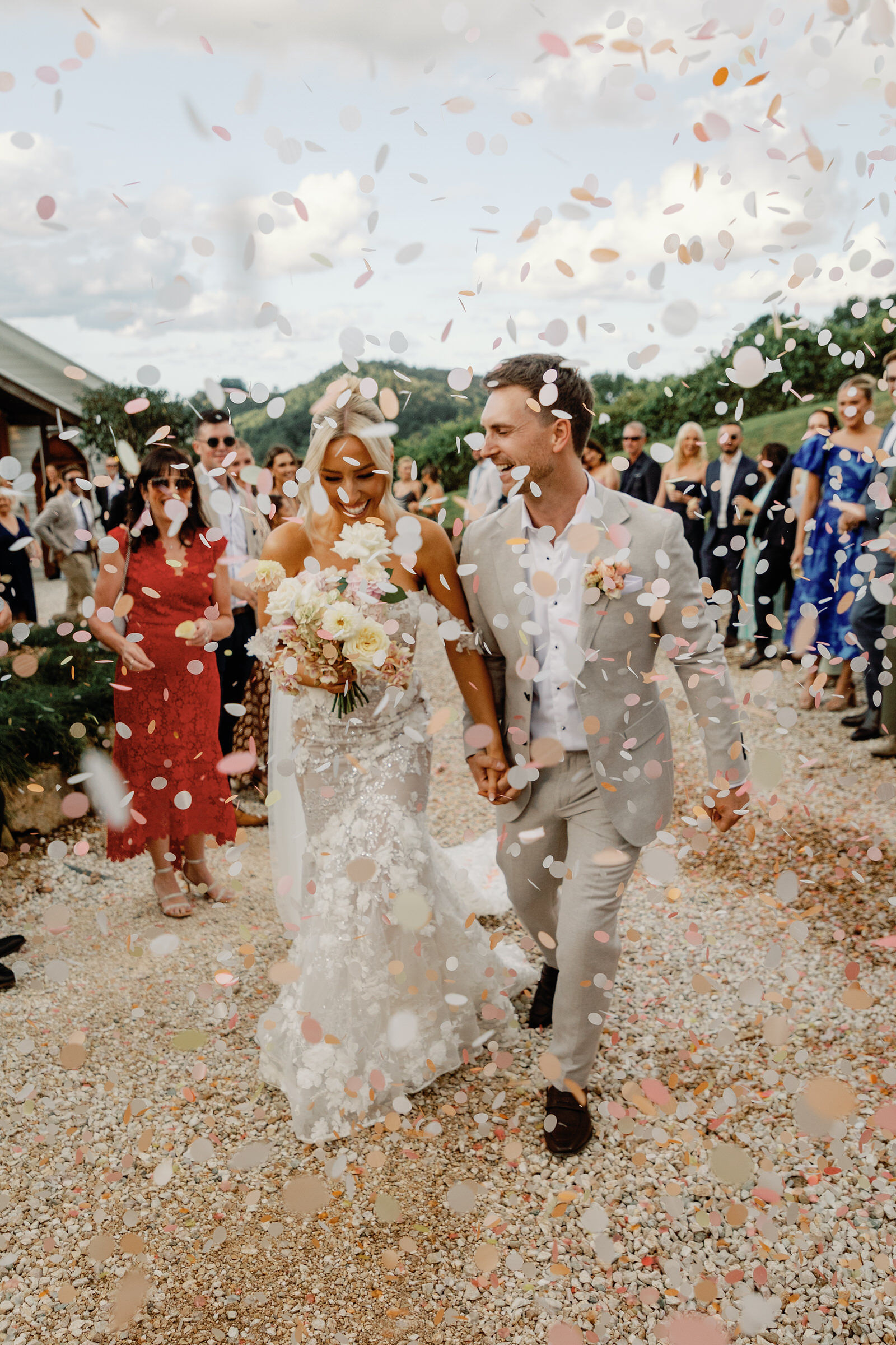 A bride and groom walk hand in hand outdoors as guests throw confetti, with smiling people in the background and a scenic view behind them.