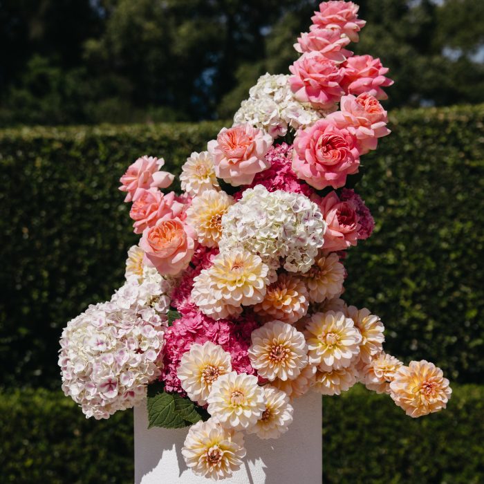 A floral arrangement of pink, peach, and cream flowers displayed on a white pedestal outdoors, with green hedges and trees in the background.