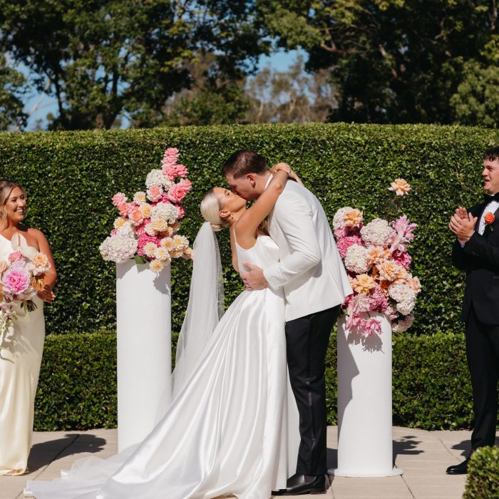 A bride and groom kiss at an outdoor wedding ceremony, surrounded by flower arrangements and attended by bridesmaids and groomsmen.