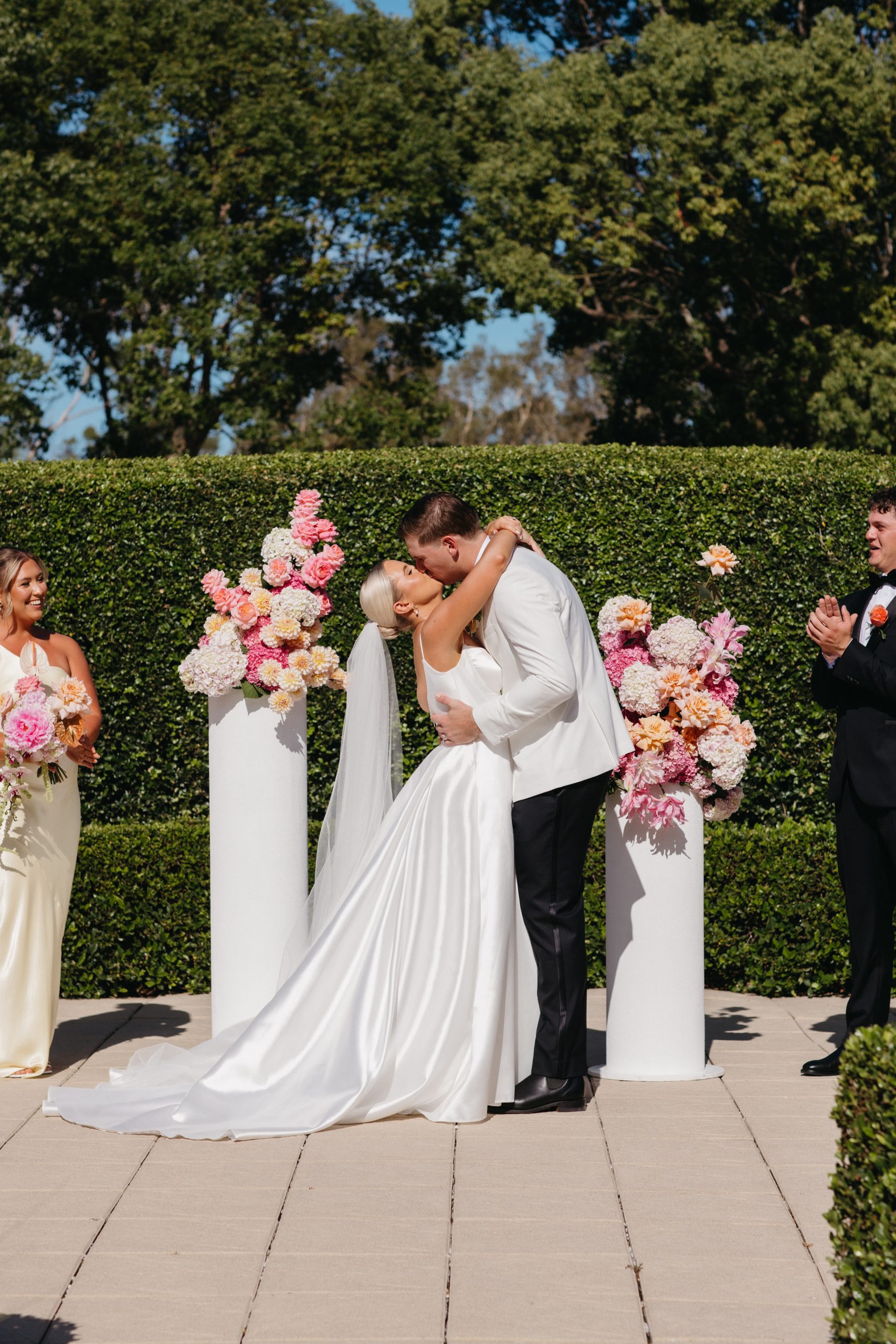 A bride and groom kiss at an outdoor wedding ceremony, surrounded by flower arrangements and attended by bridesmaids and groomsmen.