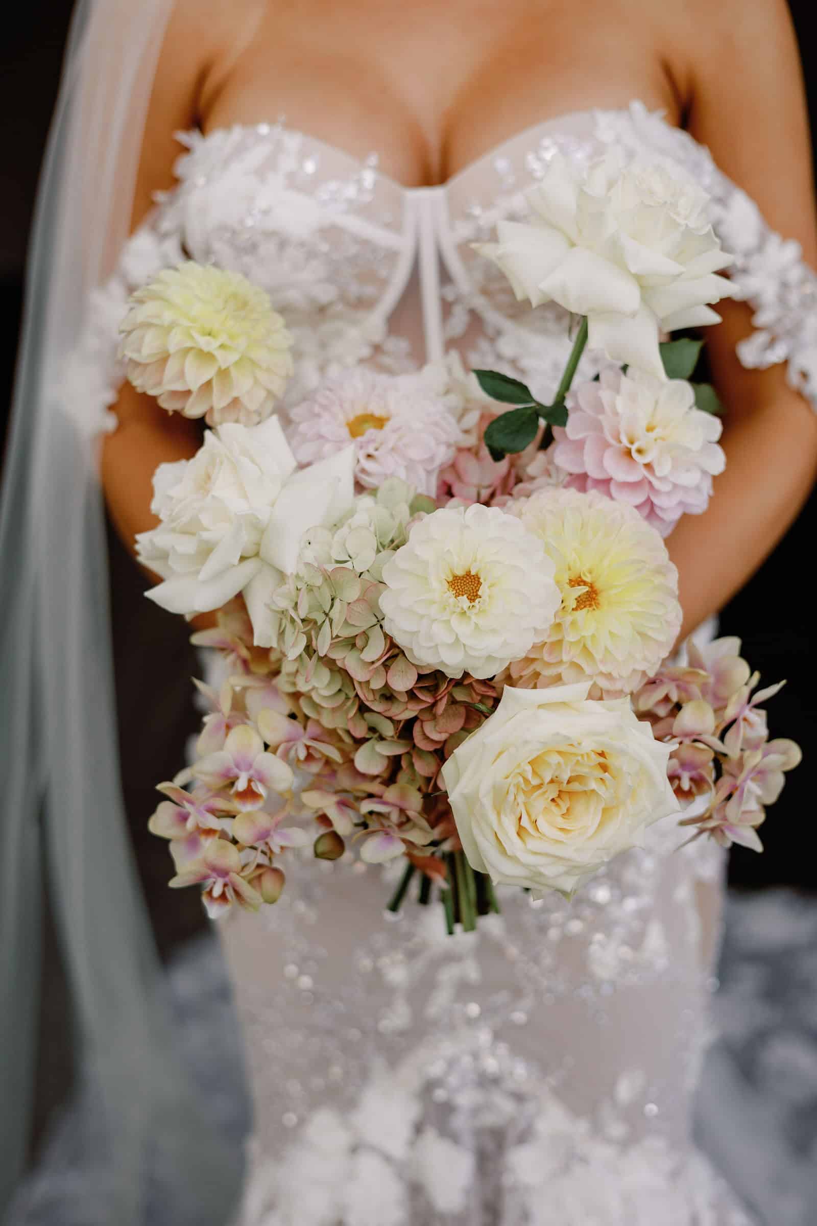 A bride in a detailed, lace wedding dress holds a bouquet of white, cream, and pale pink flowers.