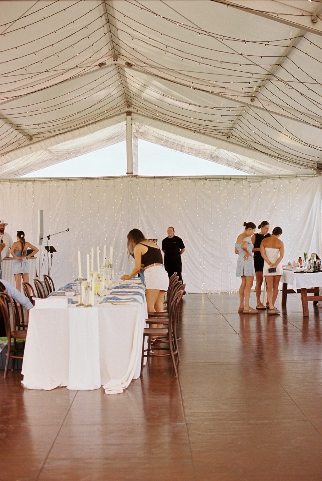 People arrange a long, decorated dining table under a large tent with string lights; several others stand and talk near a side table in the background.