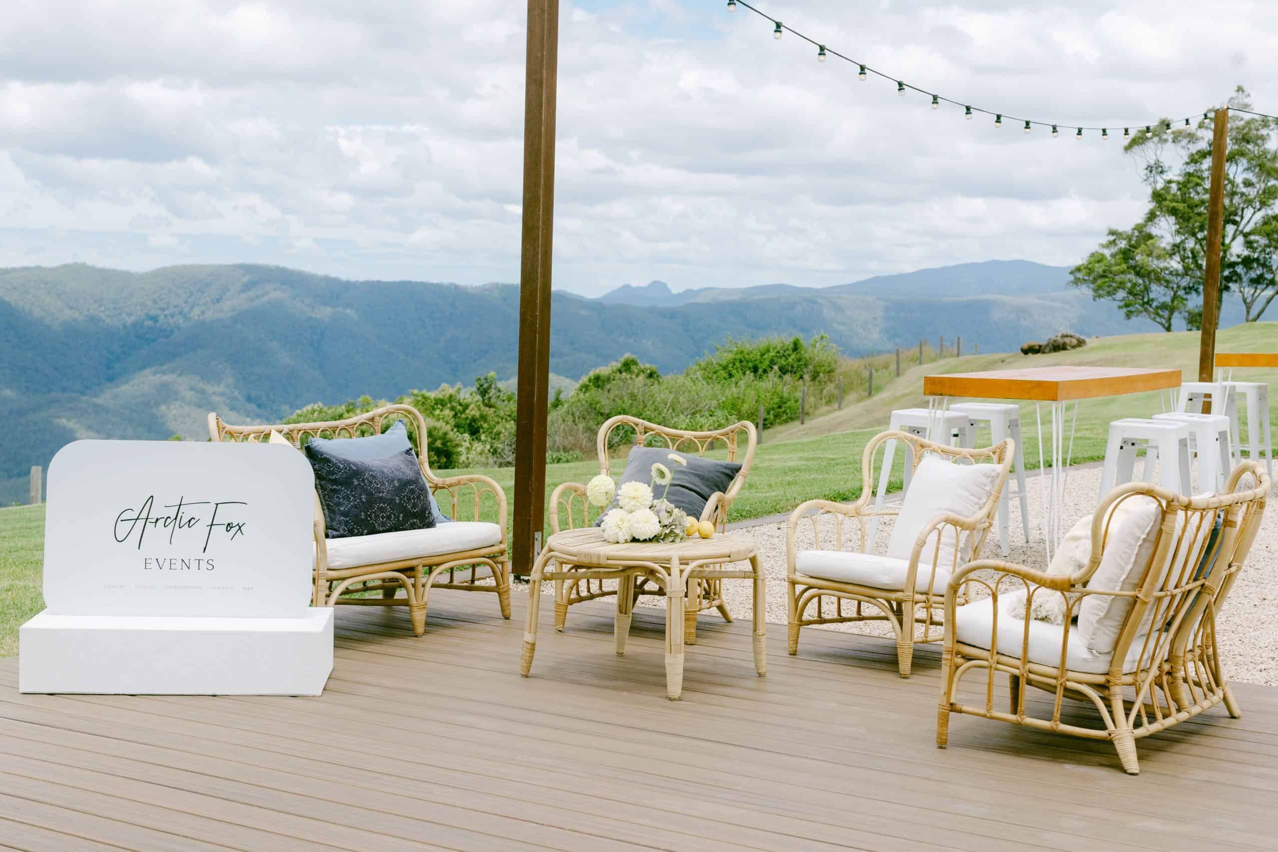Outdoor seating area with wicker chairs and a small table on a wooden deck, overlooking mountains; "Artic Fox Events" sign on the left, string lights above.