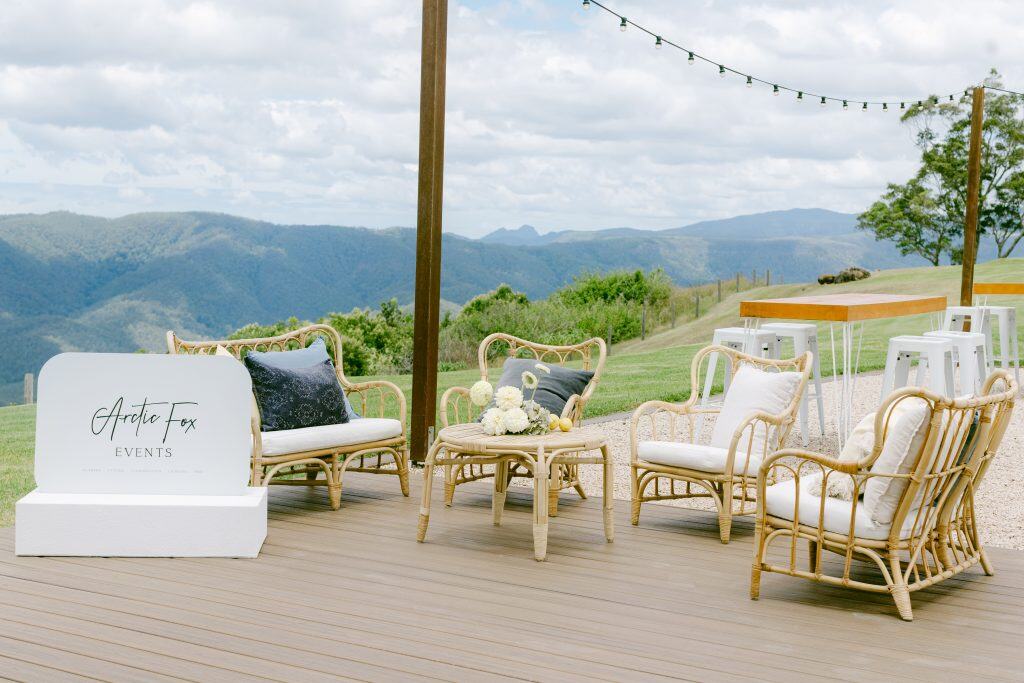 Outdoor lounge area with wicker chairs, small tables, decorative pillows, and a sign reading "Artic Fox Events," set on a wooden deck overlooking mountains and greenery.