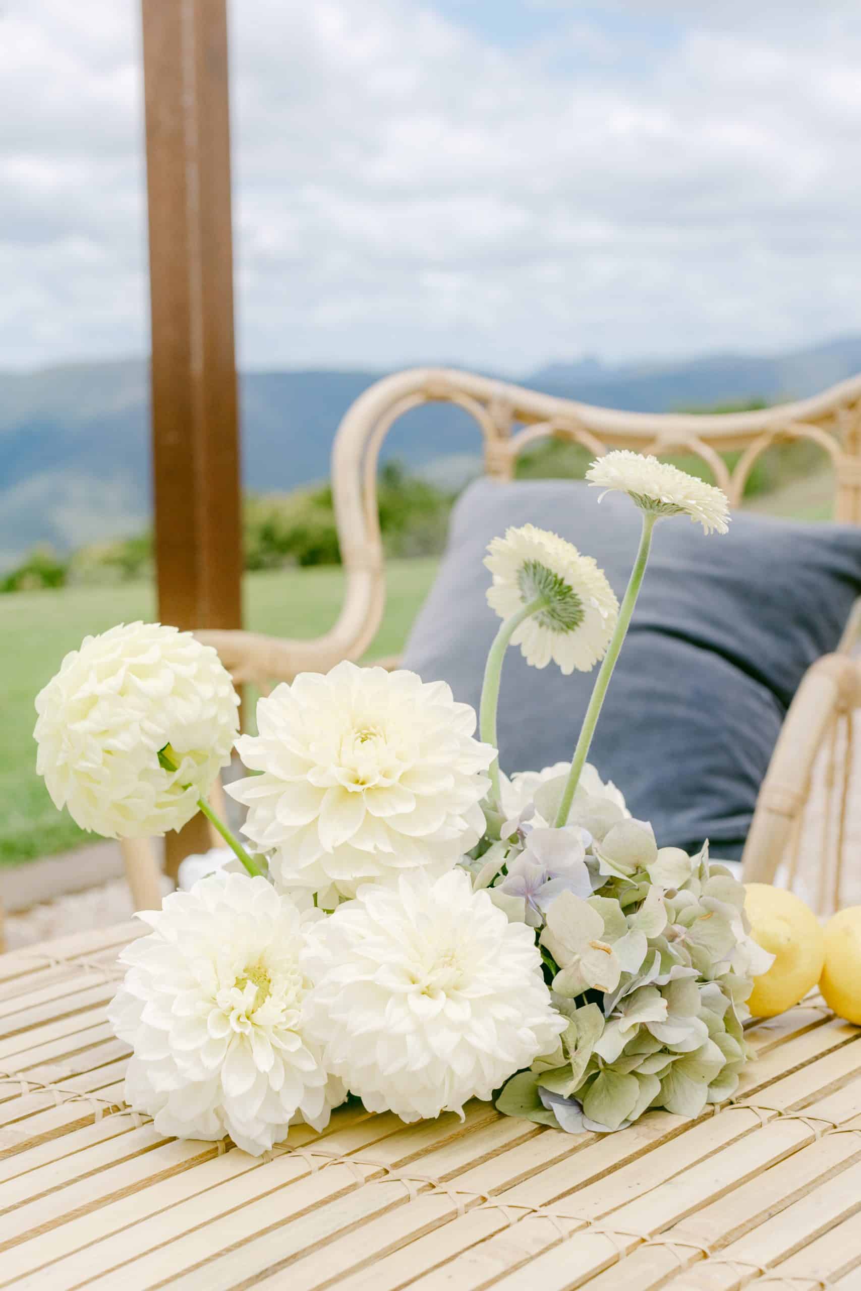 A small bouquet of white flowers rests on a bamboo table next to a wicker chair with a blue cushion, outdoors with mountains and cloudy sky in the background.