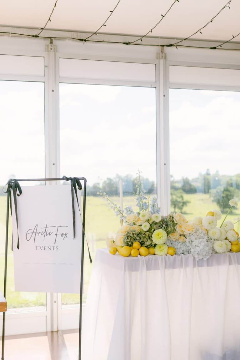 A table decorated with yellow and white flowers and lemons sits beside a white Arctic Fox Events sign in front of large windows overlooking a green landscape.